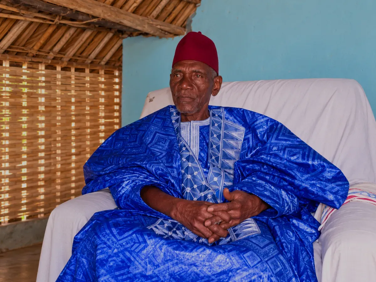 Portrait d’un marabout en grand boubou bleu brodé et chéchia rouge, assis dans un fauteuil